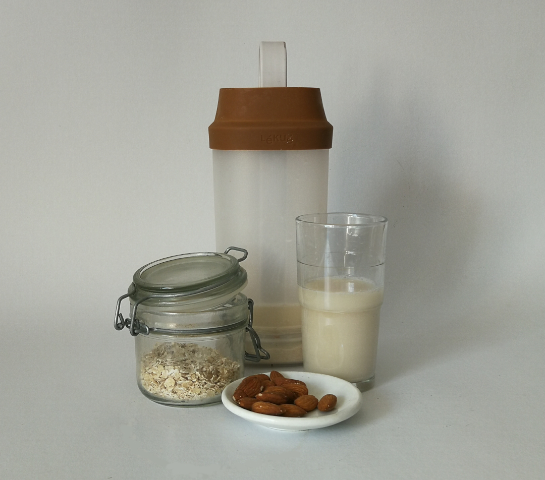 mleko roślinne, na zdjęciu pojemnik z mlekiem owsianym, szklanka z mlekiem, słoik z płatkami owsianymi, na białym talerzyku migdały