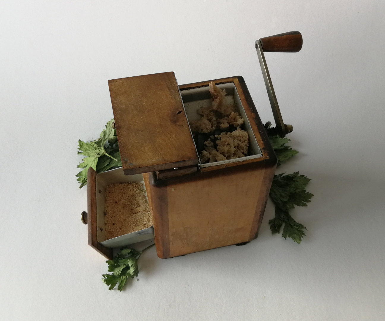 drewnianaręczna maszynka do mielenia z kawałkami pieczywa i gotową bułką tartą w uchylonej szufladzie, w tle suche listki