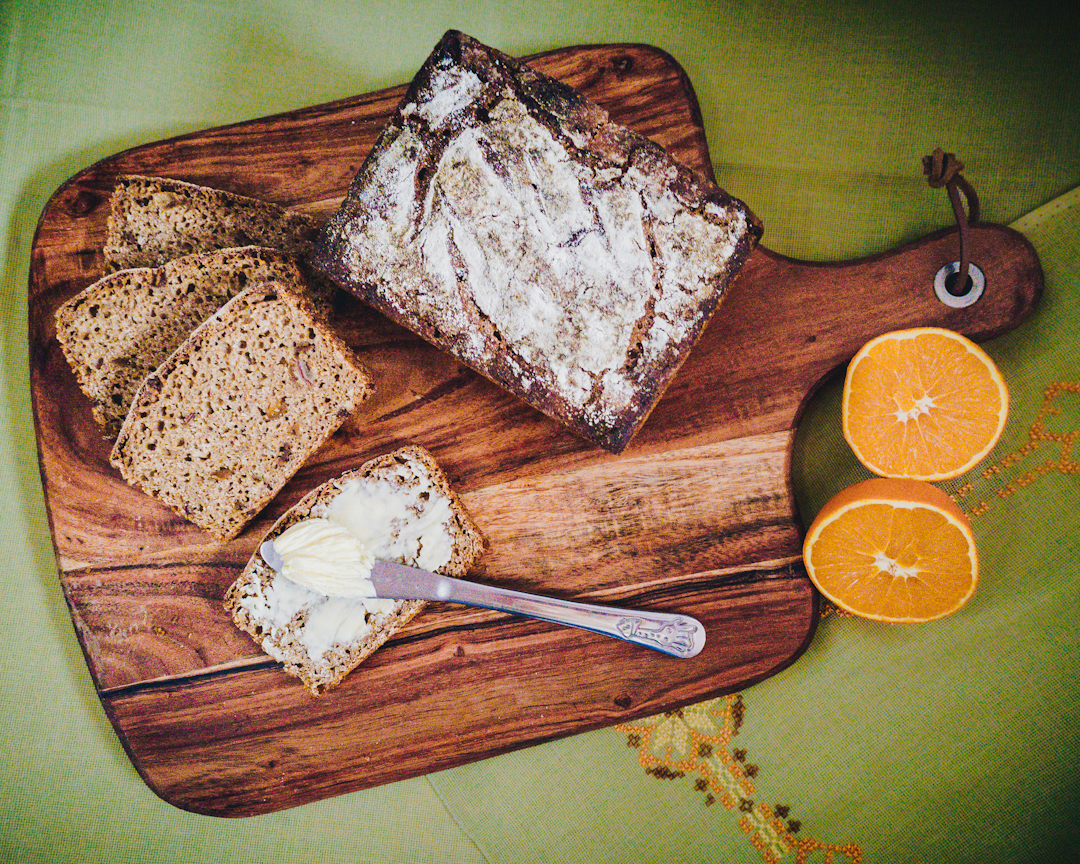 na drewnianej desce ułozone kromki chleba, w tym jedna posmarowana masłem, bochenek i dekoracja z pomarańczy
