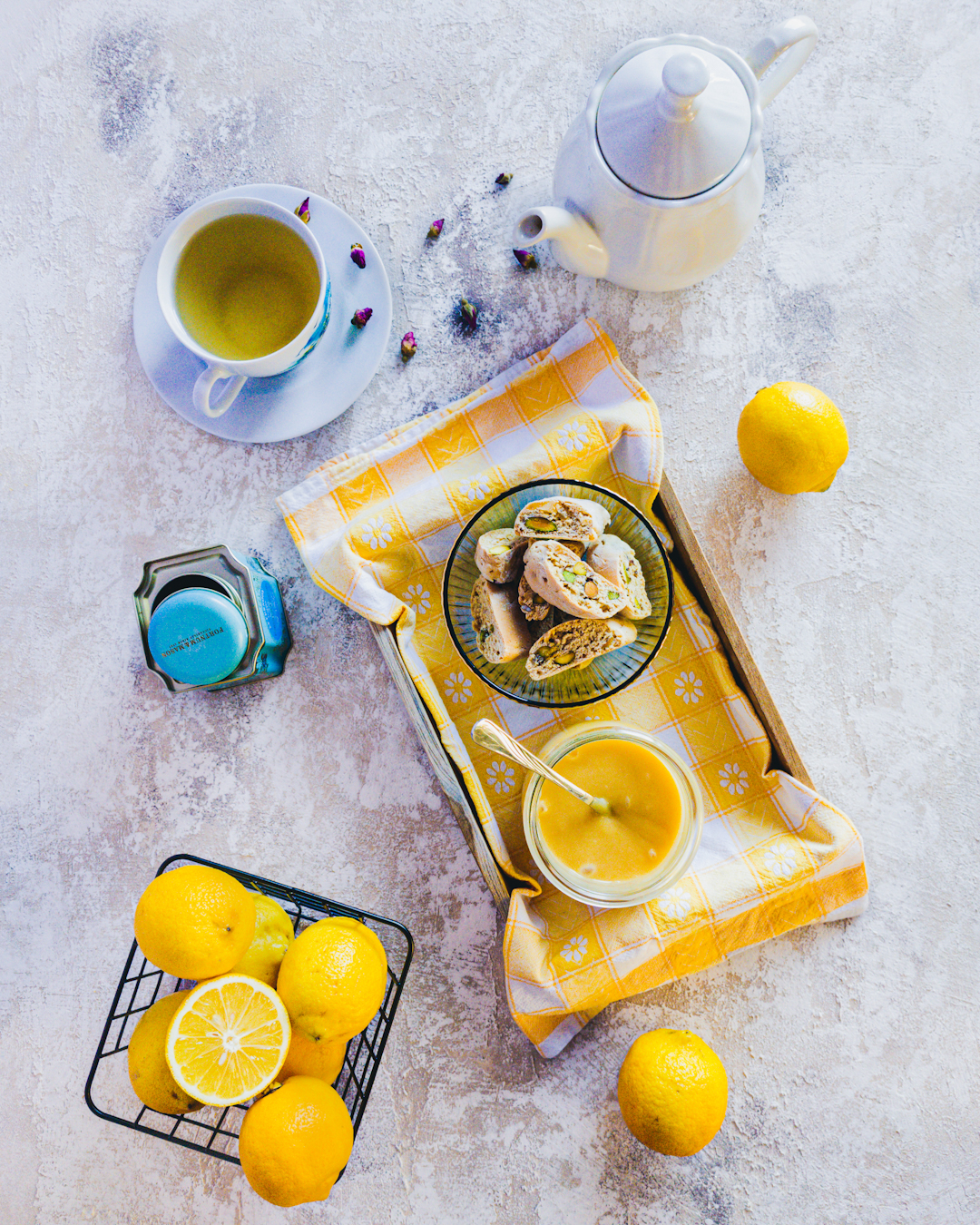Flat lay - lemon curd w towarzystwie cytryn, zielonej herbaty i cantucci. Dodatkowymi elementami dekoracji są dzbanek i puszka herbaty.