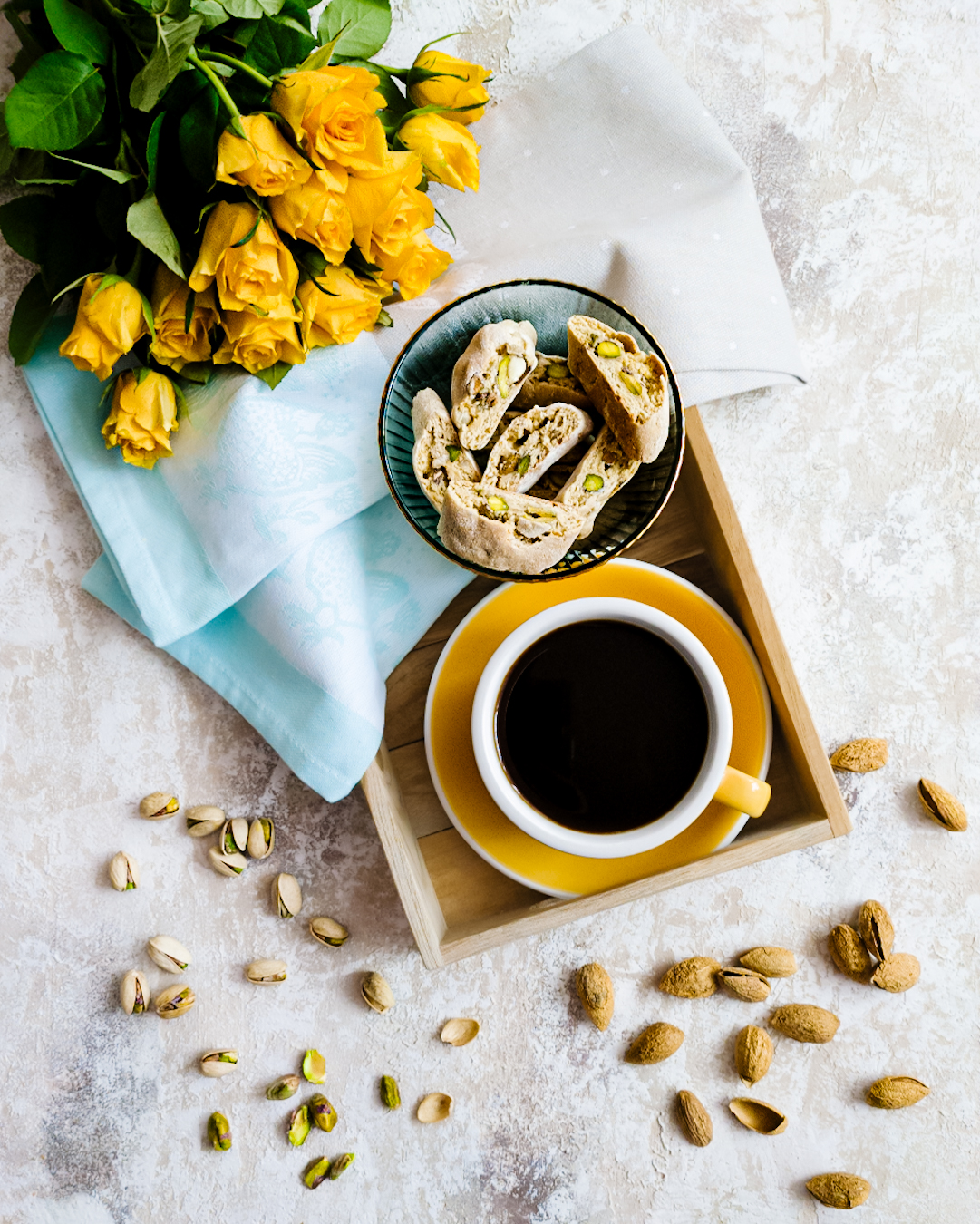 Miseczka z cantuccini w towarzystwie kawy w żółtej filiżance i żółtych róż. W tle pistacje i migdały - widok z góry.