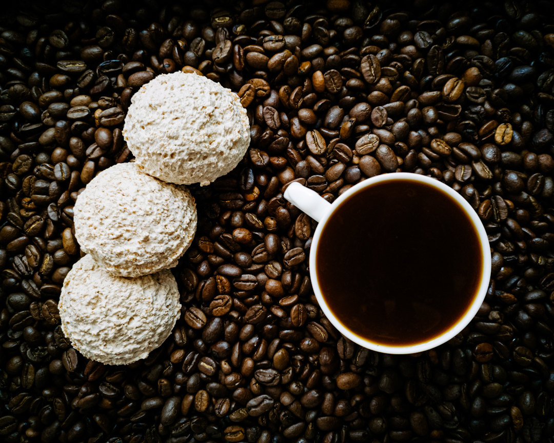 Trzy kokosanki obok filiżanka czarnej kawy, a pod nimi rozsypane ziarna kawy.