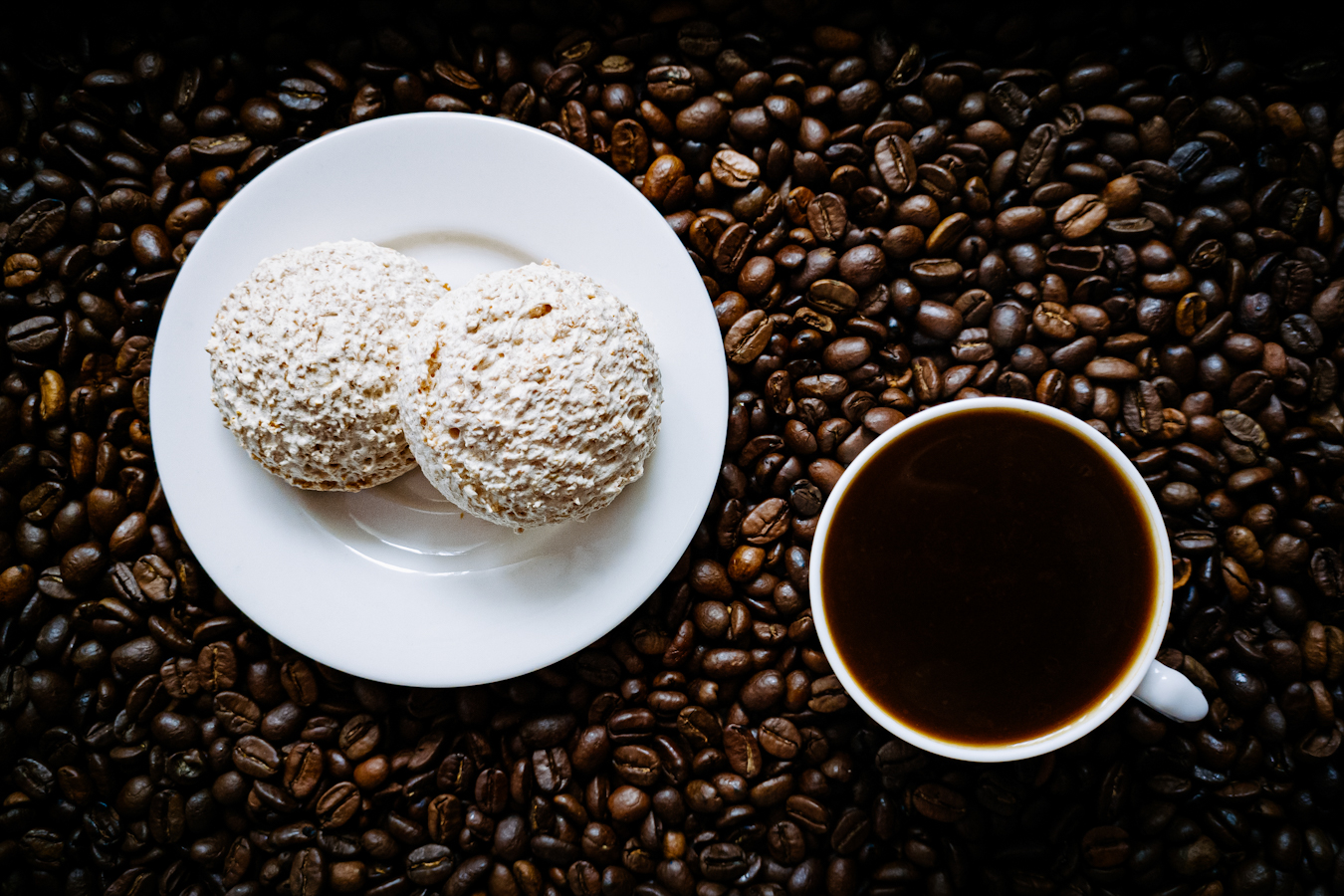 Dwie kokosanki na białym talerzyku, obok filiżanka czarnej kawy, a pod nimi rozsypane ziarna kawy.
