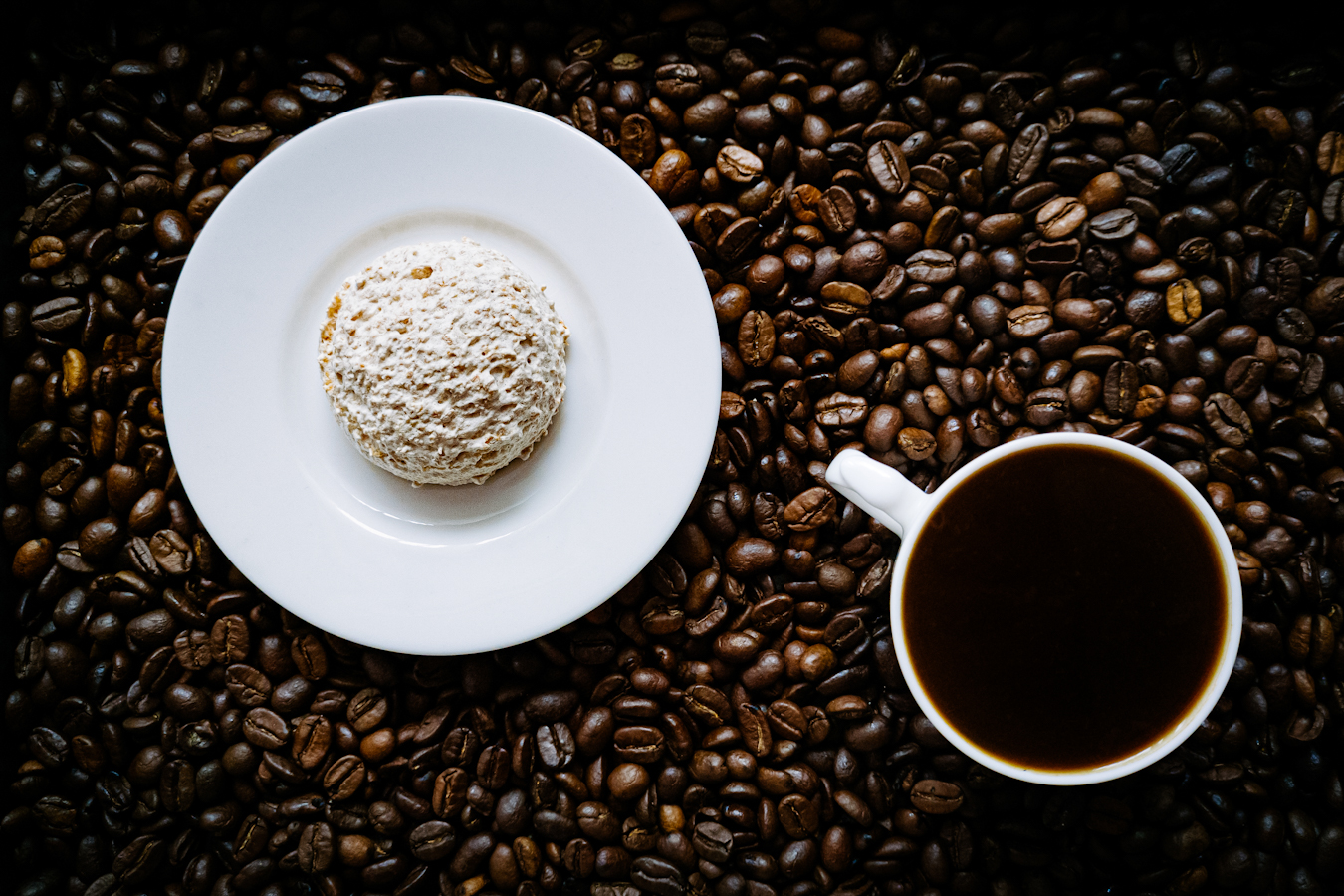 Kokosanka na białym talerzyku, obok filiżanka czarnej kawy, a pod nimi rozsypane ziarna kawy.