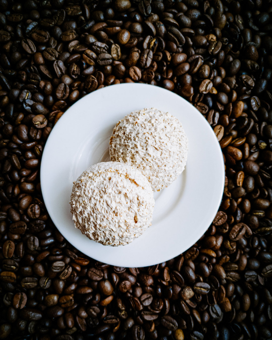 Dwie kokosanki na białym talerzyku, pod nimi rozsypane ziarna kawy.