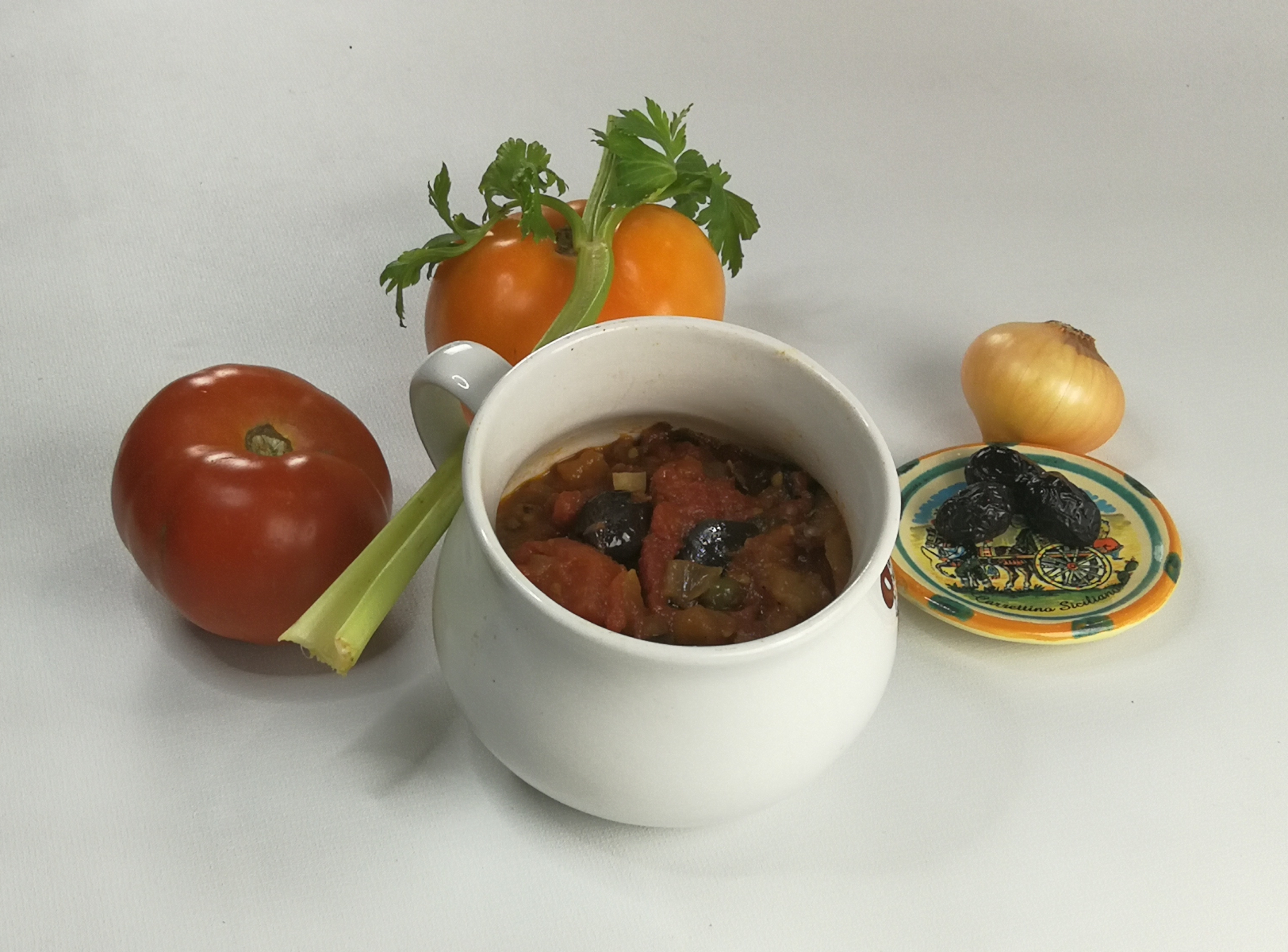 caponata w kubeczku, obok składniki: pomidory, seler naciowy, cebula oraz oliwki na talerzyku