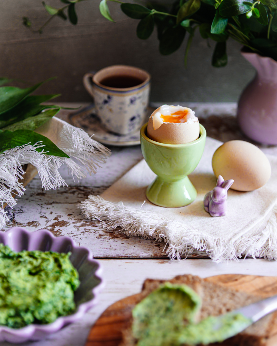 Na ściereczce stoi kieliszek z jakiem na miękko. Obok leży drugie jajko. W tle miska z zielonym masełkiem czosnkowym i filiżanka.